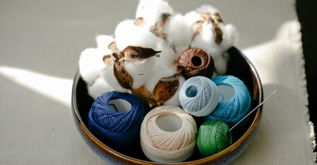 From above of sunlit clay bowl with colorful yarn for handicraft and white balls of cotton fiber placed on beige cloth on table